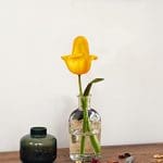 A yellow flower in a glass vase next to a green bottle.