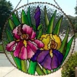 Stained glass artwork featuring colorful iris flowers in pink, purple, and yellow hues, created using the Gallery Glass Paint Most Popular Kit, displayed in a circular frame and hung against a sunny backdrop.