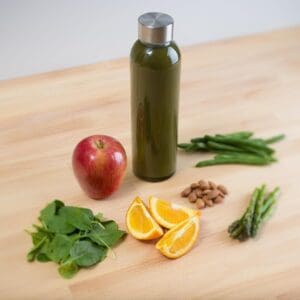 A Kombucha bottle with fresh ingredients: apple, orange slices, spinach, green beans, almonds, and asparagus on a wooden table.