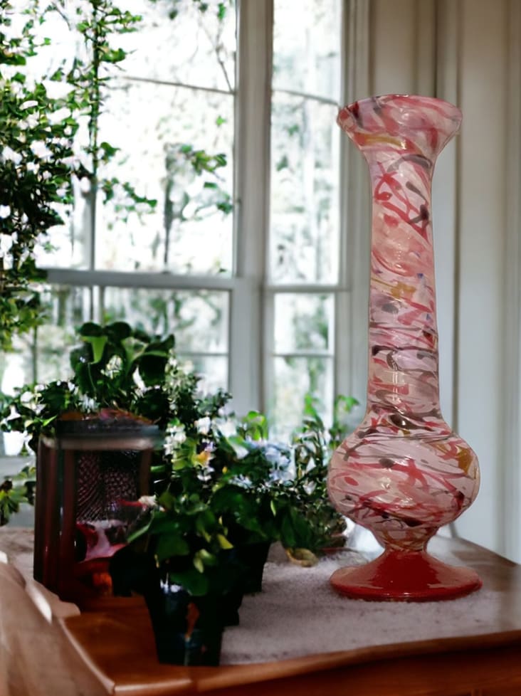 A decorative pink and white swirl glass vase on a window ledge surrounded by green plants and a lantern.