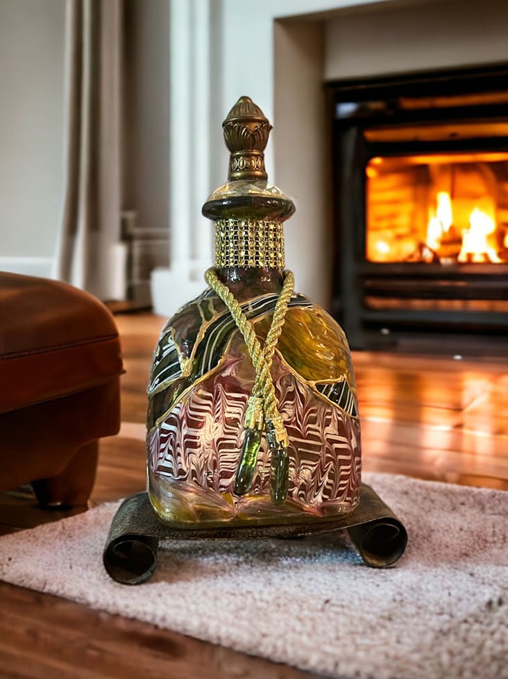 An ornate green glass decanter on a small rug with a lit fireplace in the background.