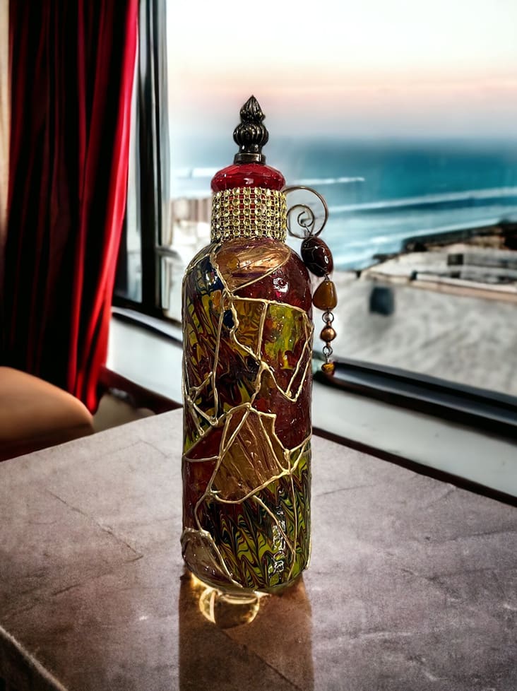 Ornate glass bottle decorated with colorful mosaic tiles and beads, placed on a table near a window overlooking the sea.