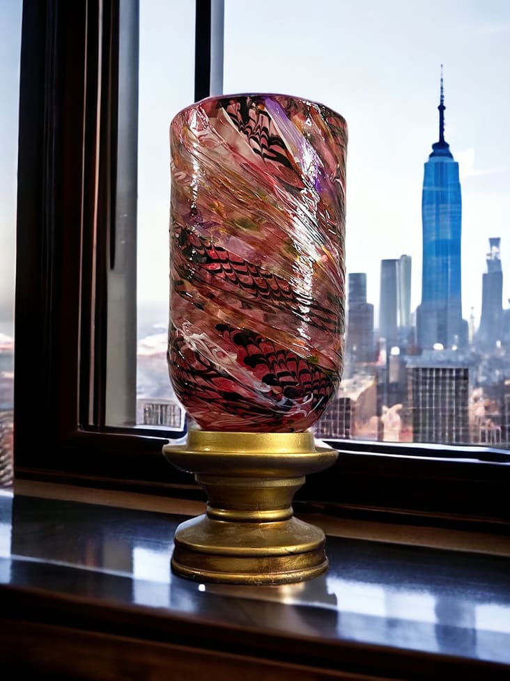 Decorative glass vase on a brass stand by a window, with a cityscape and a tall blue skyscraper in the background.
