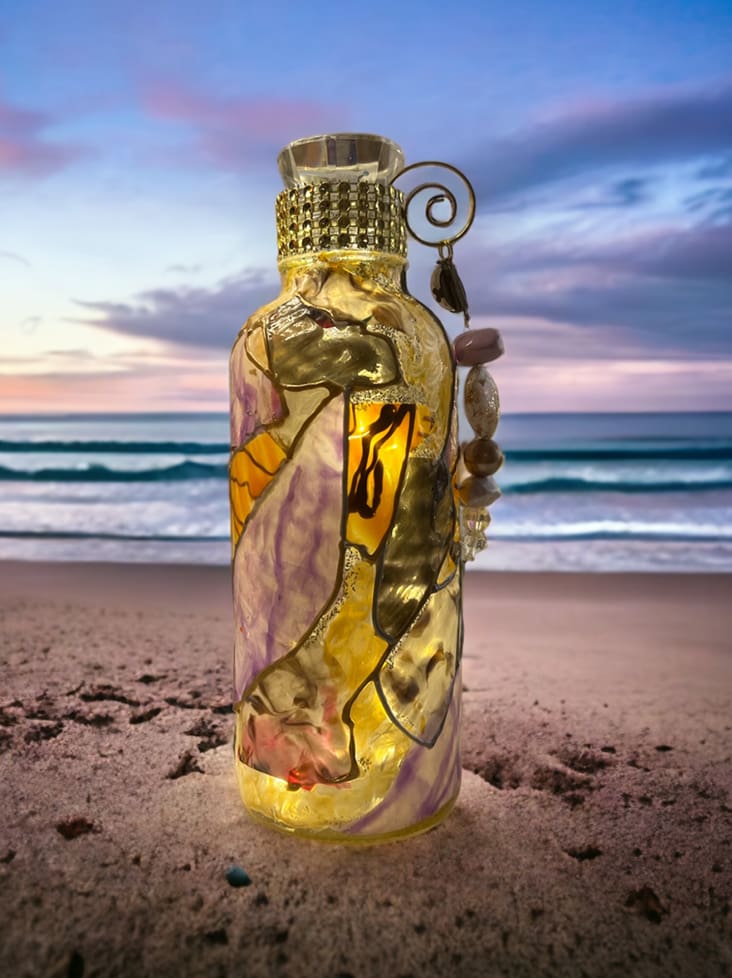 Decorative glass bottle on a beach with shells attached, against a backdrop of waves and a sunset sky.