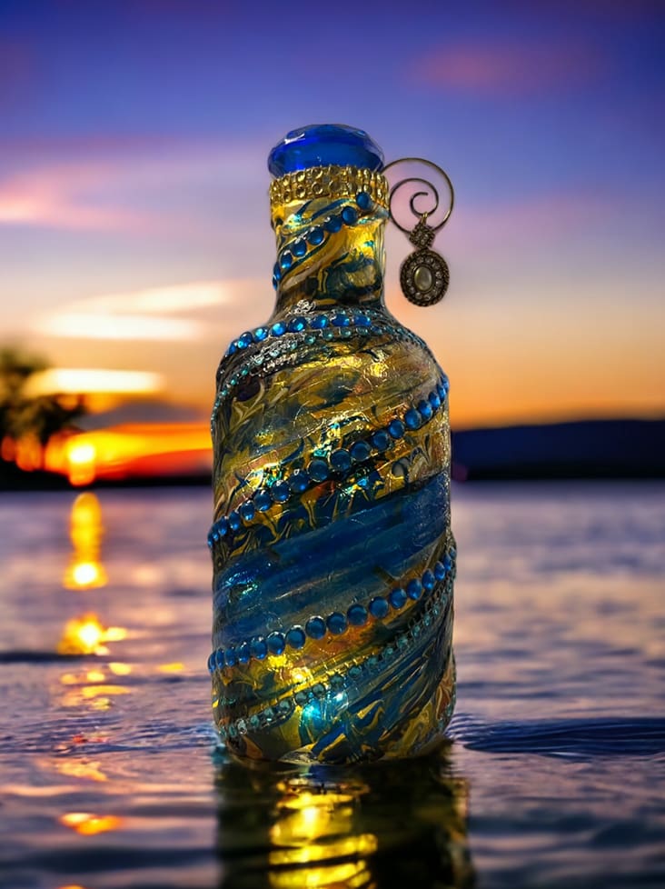 Decorative blue glass bottle with intricate patterns, partially submerged in water at sunset.