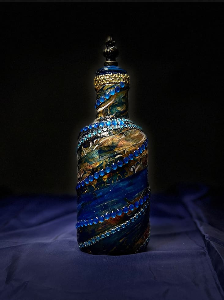 A decorative glass bottle adorned with vibrant blue and gold patterns and embellishments, highlighted against a dark background.